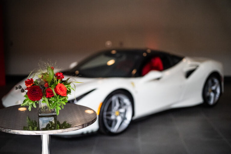 Red floral arrangement with roses, dahlias, butterfly ranunculus, hydrangea, and gardenia greens in a glass square vase, with a white Ferrari in the background. Flowers by Rebel Girl Floral.